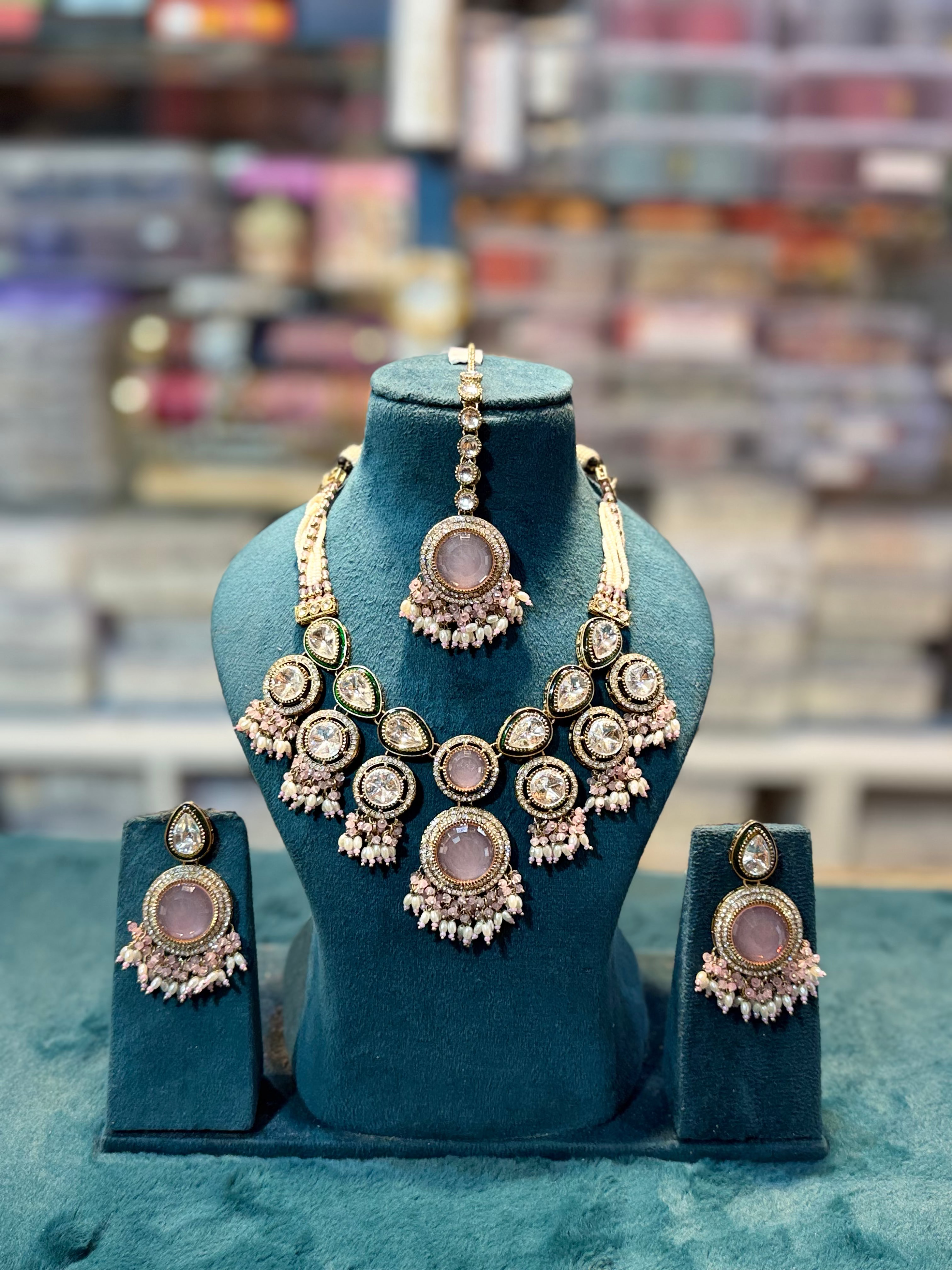 A gold-toned Indian ethnic jewelry set featuring a statement necklace, matching earrings, and a choker, displayed on a teal velvet jewelry bust. The jewelry features prominent oval pinkish-purple stones and intricate silver beadwork, set against a blurred background.